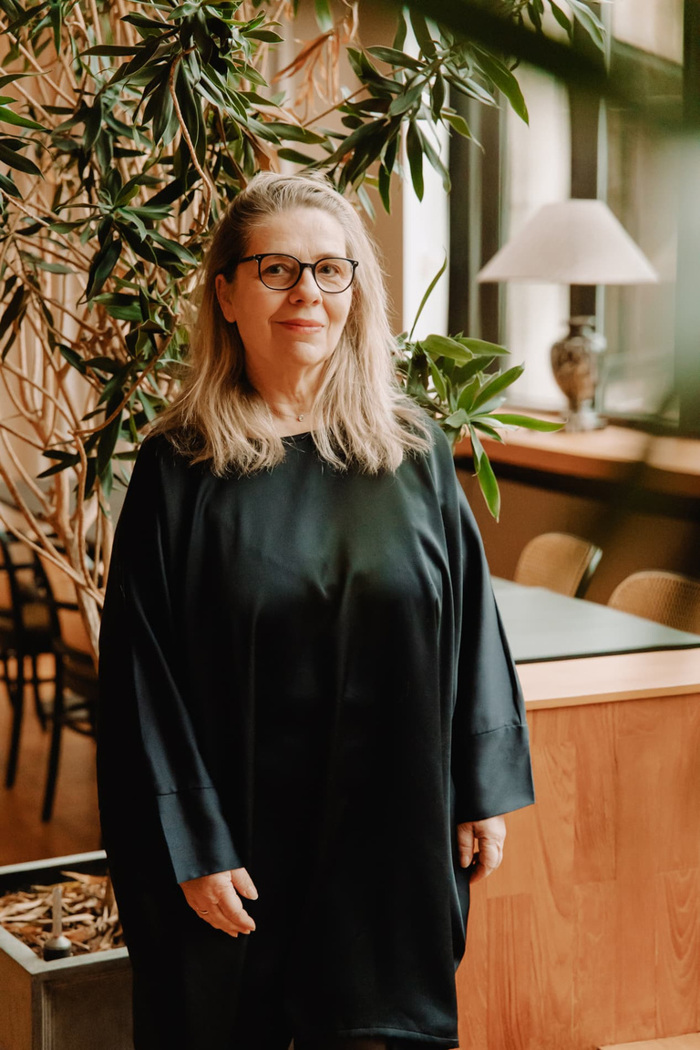 Vrouw met lange, blonde haren en bril, gekleed in een zwarte jurk, standing in een ruimte met planten en houten meubels.