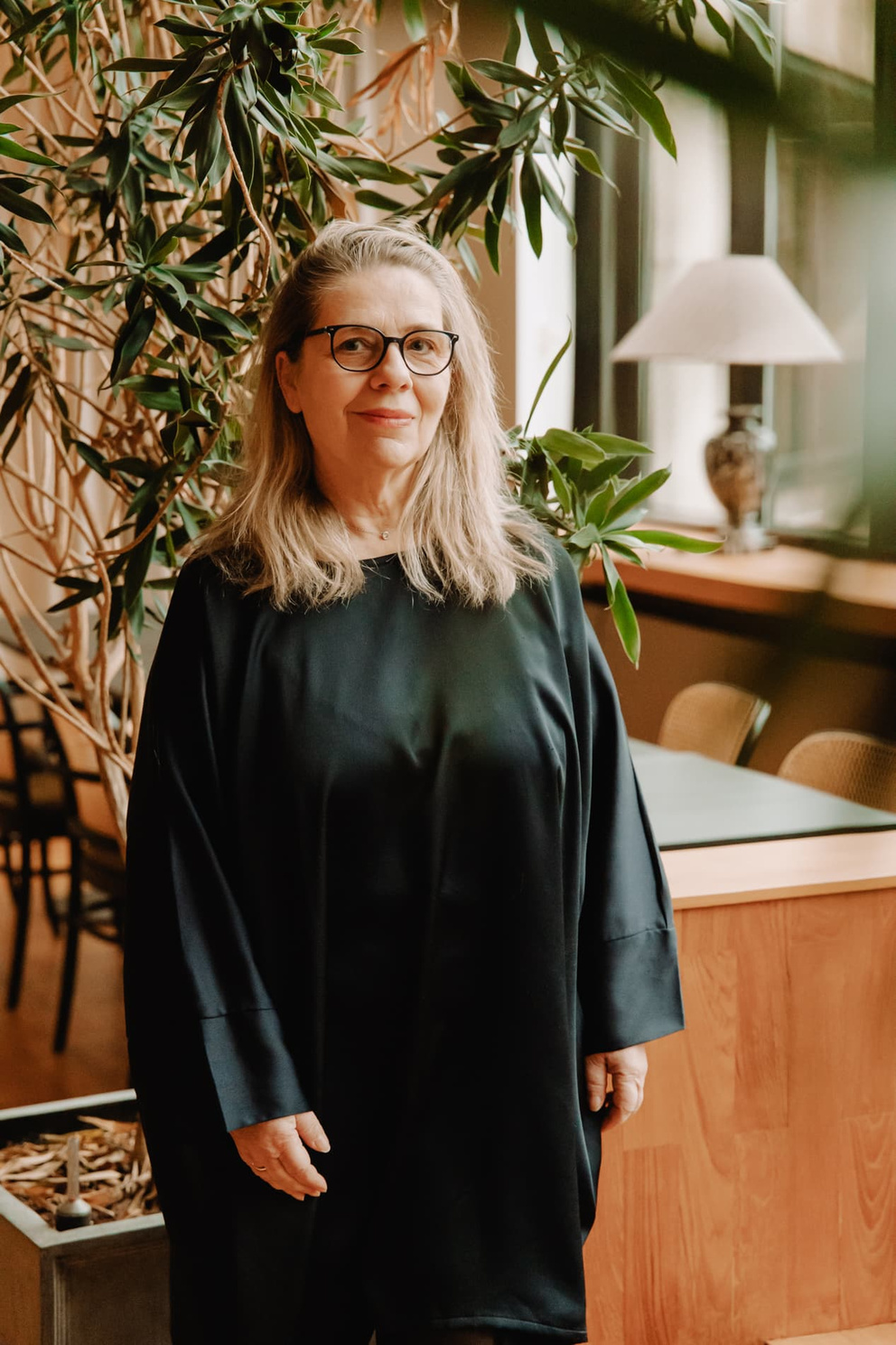 Vrouw met lange, blonde haren en bril, gekleed in een zwarte jurk, standing in een ruimte met planten en houten meubels.