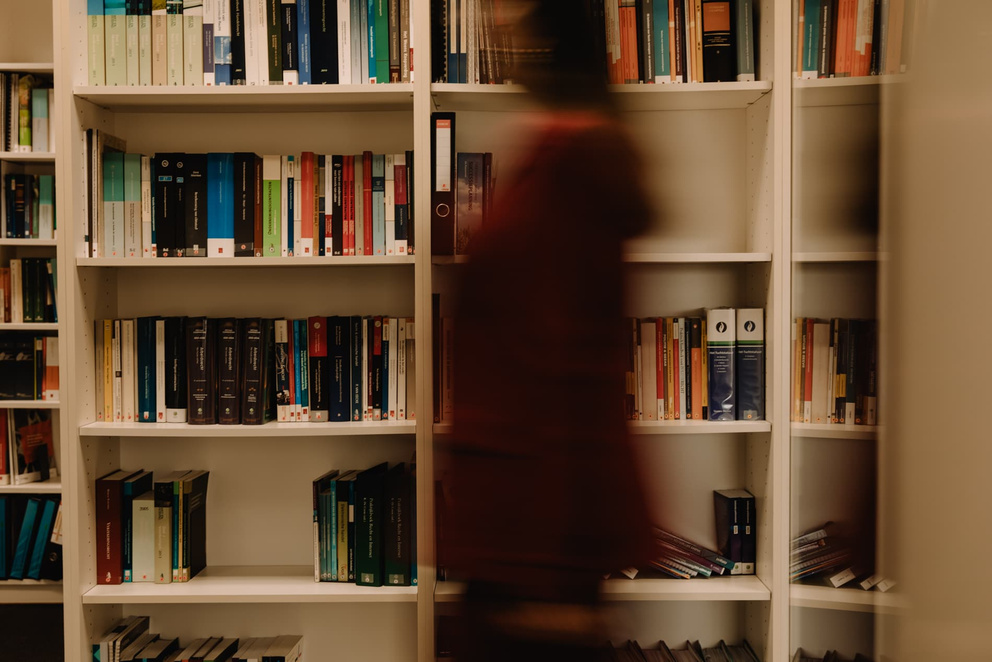 Een persoon loopt voorbij een boekenkast vol kleurrijke boeken in een bibliotheek.