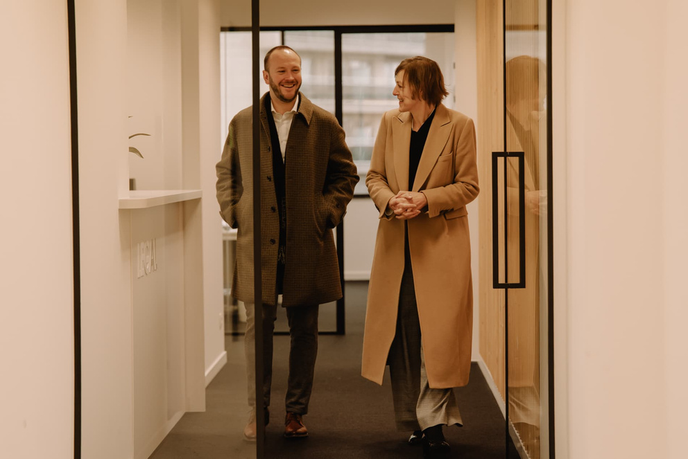Twee mensen in formele kleding lopen lachend door een moderne kantoorhal.