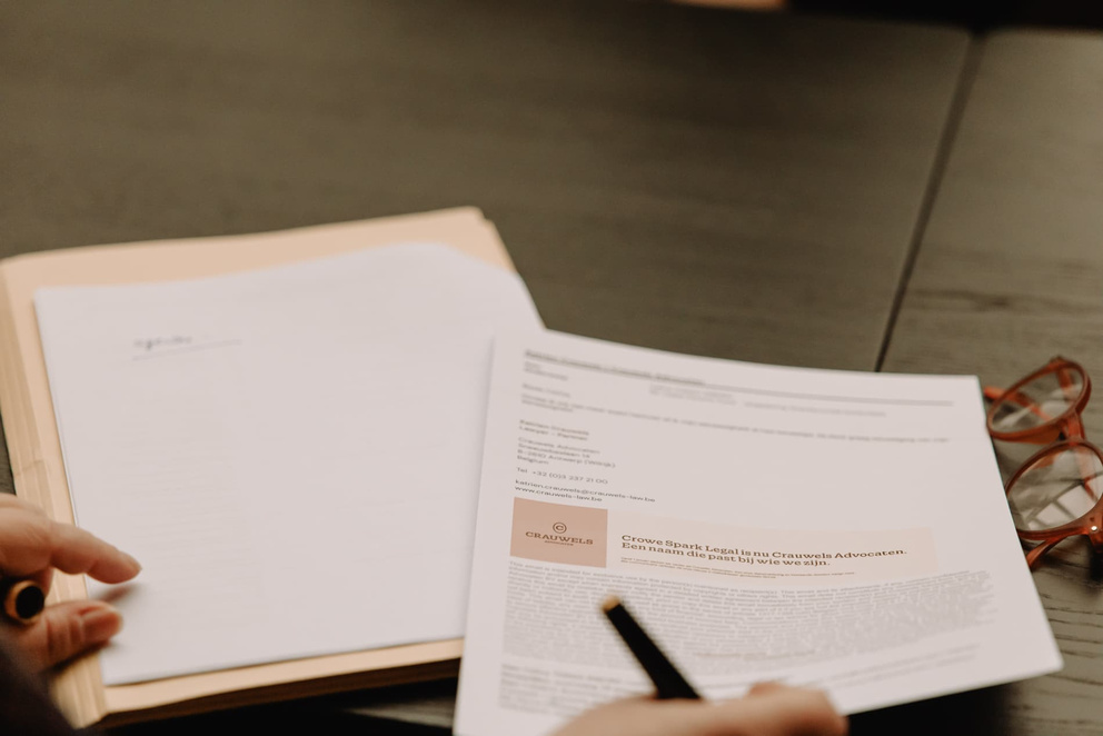 Een hand houdt een pen boven een document, met een stapel papieren en een bril op een tafel.