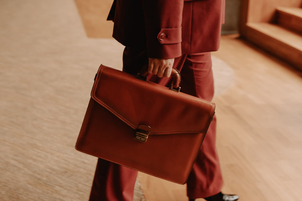 Man in een bordeauxrood pak houdt een leren aktetas vast, terwijl hij voorbijloopt in een moderne ruimte.