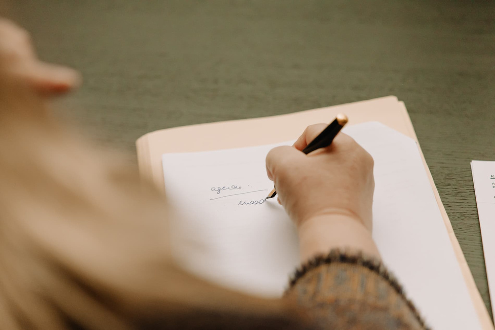 Een hand schrijft met een pen op een wit vel papier, met een stuk papier op de achtergrond.