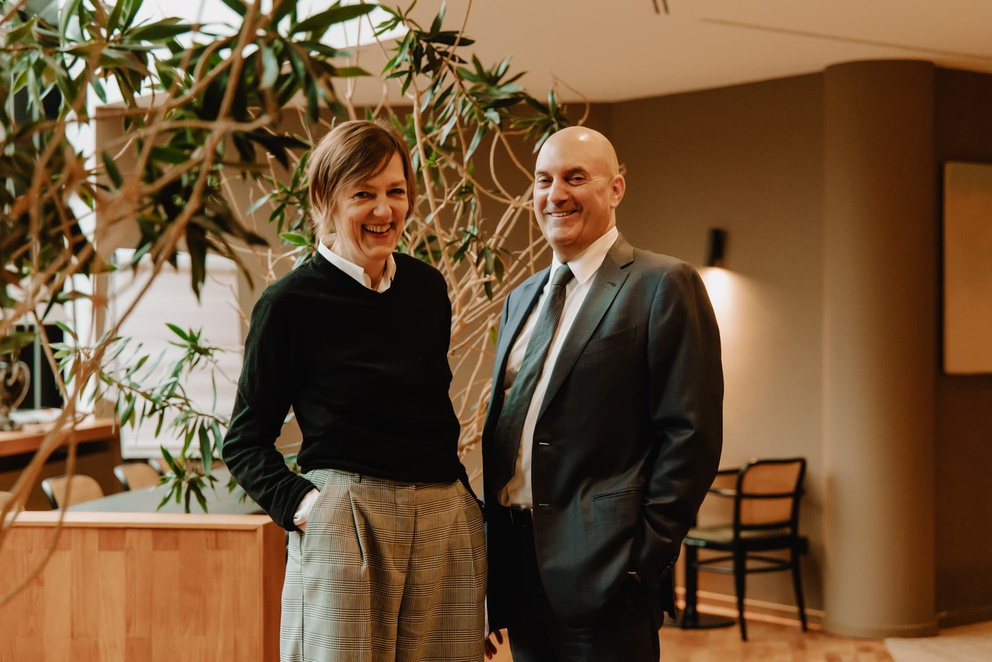 Twee professioneel geklede mensen lachen en staan naast elkaar in een moderne, groene omgeving.