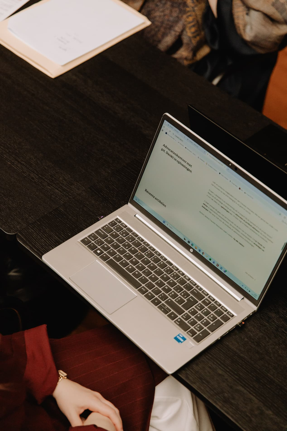 Een laptop op een tafel met een open document, met een hand in de hoek en schaduwen van mensen op de achtergrond.