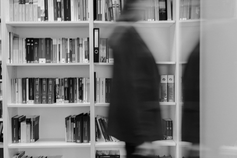 Een onscherpe figuur loopt langs een boekenplank met diverse boeken en mappen in een bibliotheek.